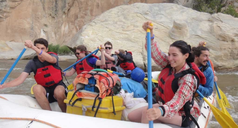 A group of students wearing safety gear paddle a green raft on a river. 